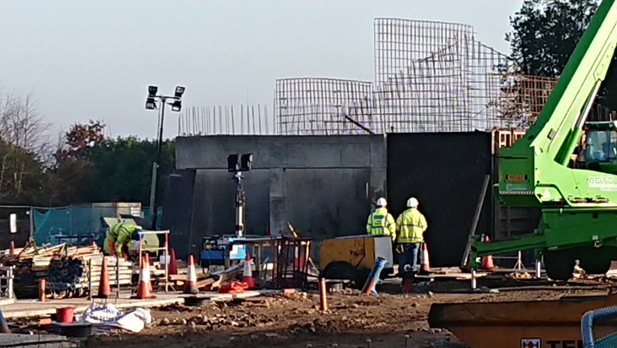Workers in high-visibility clothing on the construction site beside poured concrete enclosure walls, with steel reinforcement cages and a telehandler crane